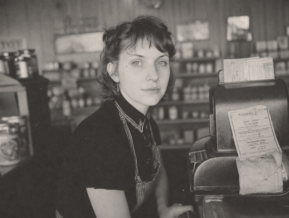 A black-and-white photograph of a woman working behind a shop counter